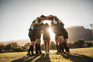 Rugby players standing in a circle with their hands on shoulders. Rugby team in huddle after the match. Bright sunshine through the huddle.